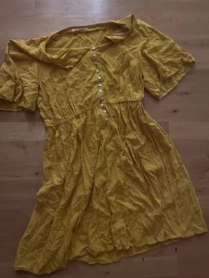 Gul kortklänning från Indiska - Säljer en söt gul klänning från Indiska i storlek M. Klänningen har korta ärmar, knappar framtill och en mjuk, rynkad midja. Materialet är lätt och känns som bomull, perfekt för varma dagar. Den har en avslappnad passform och en fin krage.