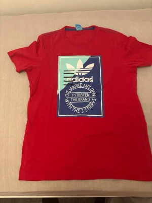 Röd Adidas t-shirt med tryck XL - Säljer en röd t-shirt från Adidas i storlek XL. Framsidan har ett stort grafiskt tryck i blått, vitt och mintgrönt med Adidas-logga och text. T-shirten har rund hals med blå kant och är kortärmad. Materialet är mjuk bomull, perfekt för en chill och sportig look.