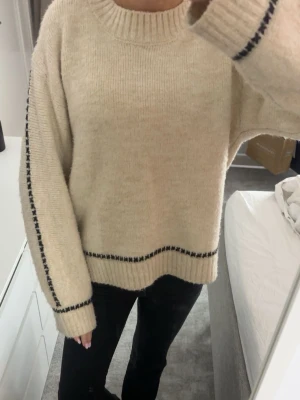 Beige stickad tröja med svarta detaljer - Säljer en beige stickad tröja med svarta korsstygn längs ärmarna och nedtill. Tröjan har en rund halsringning, ribbade muddar och en loose passform. jag har xs/s i vanliga fall och den passar perfekt!