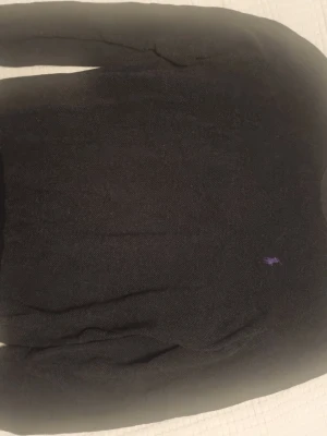 Mörkgrå stickad tröja från Polo Ralph Lauren - Klassisk mörkgrå stickad tröja från Polo Ralph Lauren med rund halsringning och lila broderad logga på bröstet. Tröjan är långärmad och gjord i mjuk bomull.