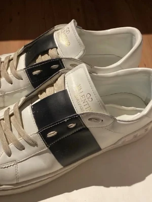Valentino Garavani vita sneakers - Säljer ett par vita Valentino Garavani sneakers med bred svart läderdetalj och silverfärgade nitar på sidan. Skorna har beige snörning och klassisk rund tå. Kommer med originalkartong och påsar. Perfekta för dig som gillar exklusiva och stilrena sneakers.