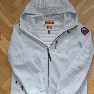 Vit jacka med luva från Parajumpers - Säljer en ljusblå jacka från Parajumpers med huva och quiltade partier på bröst och rygg. Jackan har dragkedja framtill, en bröstficka med dragkedja och en patch på ärmen. Perfekt för kyliga dagar.