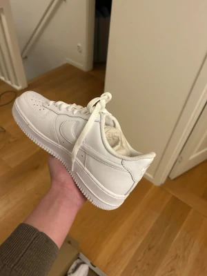 Nike Air Force 1 helvita sneakers - Säljer ett par klassiska Nike Air Force 1 i helvitt skinn. Skorna har rund tå, perforerad tåbox och vit sula. De har snörning och den ikoniska swoosh-loggan på sidan. Kommer i originalkartong. Perfekta för dig som vill ha en clean och tidlös sneaker. Skorna är sprillans nya och har aldrig använts, vann de i en tävling och storleken var för liten 