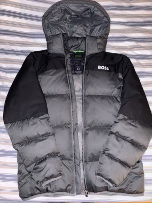 Svart och grå pufferjacka från BOSS - Säljer en stilren svart och grå pufferjacka från BOSS med huva och dragkedja. Jackan har dunfyllning, vattenavvisande material och lättviktigt tyg. BOSS-logga på bröstet och insidan. Perfekt för kalla dagar när du vill hålla värmen och ändå se snygg ut. Jackan är helt oanvänd och blev köpt i fel storlek. Kvitto finns och pris kan diskuteras!