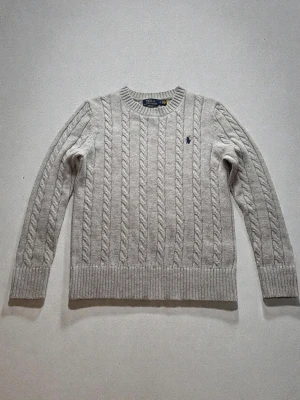 Grå kabelstickad tröja Polo Ralph Lauren - Kabelstickad grå tröja från Polo Ralph Lauren med rund halsringning och svart logga på bröstet. Tröjan har ribbade muddar och är långärmad, perfekt för kyliga dagar.