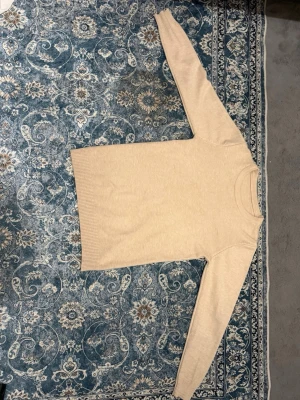 Beige stickad tröja med rund hals - Säljer en enkel och stilren beige stickad tröja med rund halsringning. Tröjan har långa ärmar och ribbade muddar vid ärmslut och nederkant. Perfekt för lager på lager under kyliga dagar.