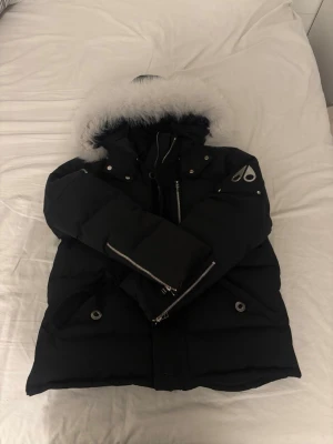 Moose Knuckles jacka - Säljer en svart pufferjacka från Moose Knuckles med fluffig vit pälskant på huvan och silverfärgade detaljer.  Inga skador eller hål i väldigt bra skick!