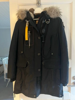 Svart Parajumpers parkas med pälskrage - Säljer en svart Parajumpers parkas med stor luva och pälskant. Jackan har flera fickor, gul detaljrem och robusta knappar samt dragkedja. Perfekt för kalla dagar och riktigt snygg streetstil. Materialet är polyester och päls.