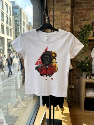 Vit Squid Game t-shirt med tryck - Vit t-shirt med färgstarkt tryck inspirerat av Squid Game. Motivet visar flera karaktärer och symboler från serien i rött, svart och gult. T-shirten har rund hals och korta ärmar, tillverkad i mjuk bomull och har en normal passform.