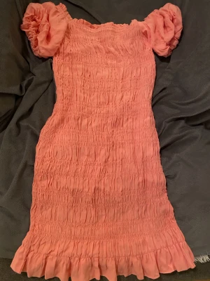 Rosa kortklänning med puffärmar XS - Supersöt rosa kortklänning med smockad struktur och volangkant nertill. Klänningen har puffiga korta ärmar och är gjord i ett lätt och mjukt tyg, perfekt för varma dagar. Offshoulder-modell som ger en snygg detalj 