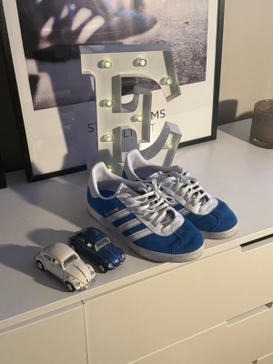 Adidas Real Madrid Gazelle blå/vit - Säljer ett par Adidas Gazelle Real Madrid edition i blå mocka med vita detaljer och klassiska tre ränder. Skorna har vit platt sula, snörning och Real Madrid-logga på hälen. Perfekt för dig som vill sticka ut med en sportig och stilren sneaker. ANVÄNDA 2 gången