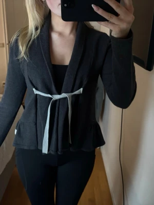 Mörkgrå kofta från Odd Molly med knyt - Säljer en mörkgrå kofta från Odd Molly med ljusgrå knytband i midjan. Koftan har lång ärm, volangkant nedtill och är tillverkad i mjukt material som känns riktigt skönt. Perfekt för lager på lager och snygg till både jeans och tights.