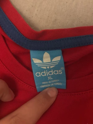 Röd Adidas t-shirt med tryck XL - Röd t-shirt från Adidas i storlek XL med blå detaljer vid kragen och stort grafiskt tryck framtill i blått, vitt och mint. Klassisk rund hals och korta ärmar. Perfekt för dig som gillar sportig och street-inspirerad stil.