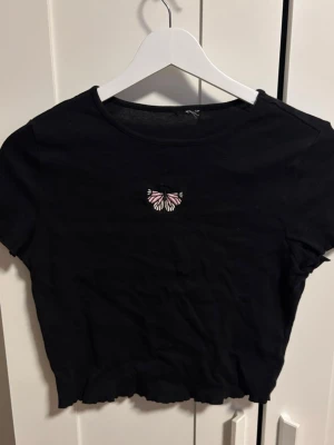 Svart croppad t-shirt med fjäril - Svart croppad t-shirt med korta ärmar och en liten broderad fjäril i rosa och vitt på bröstet. T-shirten har vågig kant nertill för en extra detalj. Storlek står inte men skulle säga S/M