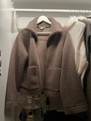 Brun teddyjacka med dragkedja - Mysig brun teddyjacka med hög krage och stora fickor framtill. Jackan har en avslappnad passform och stängs med dragkedja. Perfekt för kyliga dagar när du vill vara både varm och snygg.