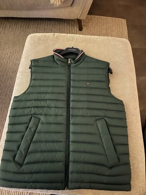 Grön dunväst från Tommy Hilfiger - Säljer en mörkgrön dunväst från Tommy Hilfiger med quiltad design och dragkedja framtill. Västen har två snedställda fickor och en hög krage med diskreta rödvita detaljer. Perfekt för lager-på-lager och snygg till jeans eller hoodie.