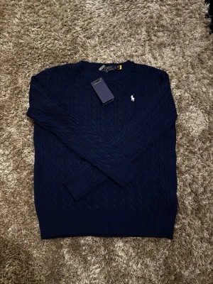 Marinblå tröja Polo Ralph Lauren - Säljer en marinblå kabelstickad tröja från Polo Ralph Lauren med klassisk rund hals och vit broderad logga på bröstet. Tröjan har lång ärm och är gjord i mjuk bomull, perfekt för lager-på-lager under kyliga dagar.