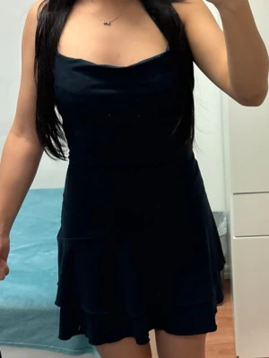 Marinblå kortklänning - Säljer en marinblå kortklänning med dubbla volanglager i kjolen. Klänningen har en snygg, enkel design med lätt draperad urringning och öppen rygg. Perfekt för dig som vill ha en stilren och chic look.