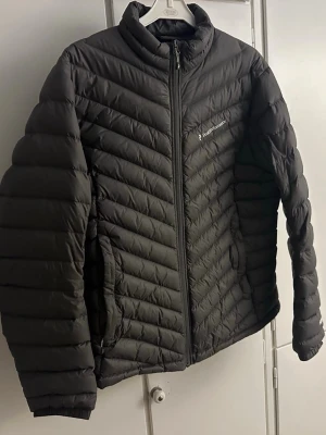 Svart dunjacka Peak Performance dam XL - Säljer en svart dunjacka från Peak Performance i dammodell, storlek XL. Jackan har quiltad design med horisontella och snedställda sömmar, hög krage och dragkedja framtill. Perfekt för kyliga dagar och riktigt snygg till alla outfits.