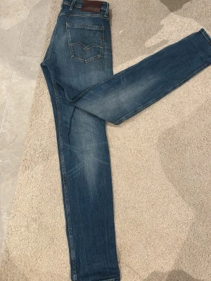 Replay Anbass jeans blå, storlek 28 - Säljer ett par snygga Replay Anbass jeans i klassisk blå tvätt. Modellen har fem fickor, normal passform och raka ben. Jeansen har diskreta slitningar och Replay-logga på fickan. Perfekta till en avslappnad stil och riktigt sköna att bära.