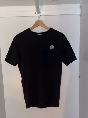 Svart Moncler t-shirt  - Svart t-shirt från Moncler med klassisk rund hals och diskret logotyp-patch på bröstet. Tillverkad i mjuk bomull för en skön känsla och enkel stil. Perfekt till jeans eller shorts för en clean look.