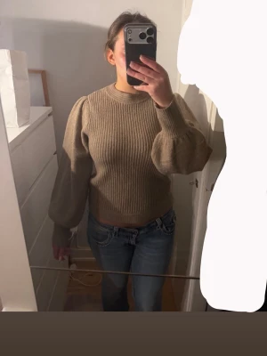 Beige stickad tröja med puffärm - Säljer en beige stickad tröja med ribbat mönster och snygga puffärmar. Tröjan har rund halsringning och är lite croppad i modellen. Perfekt för dig som vill ha en trendig och mysig look.