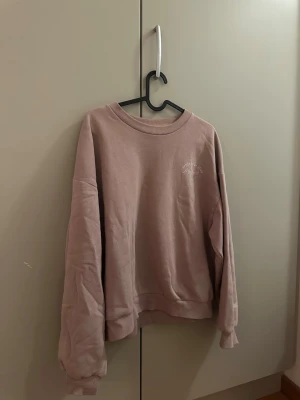 Lila collagetröja - Säljer en lila collagetröja med broderad logga på bröstet. Tröjan har rund hals, ribbade muddar och är långärmad. Perfekt för chill dagar och enkel att matcha med jeans eller mjukisbyxor.