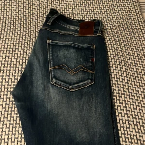Replay Anbass blå jeans, midwaist, W30 L32 - Snygga Replay Anbass jeans i blå tvätt med slitningar och trasiga detaljer. Klassisk femficksmodell med coola kontrastsömmar på bakfickan. Midwaist passform och tillverkade i stretchig jeanstyg för extra komfort. Perfekta för en avslappnad streetstil.