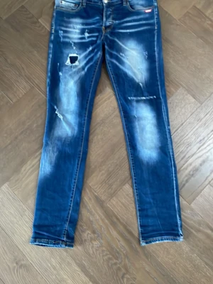 Blå distressed slim fit jeans från Dsquared2💰 - Säljer ett par blå jeans från Dsquared2 med slitna detaljer och patch på vänster lår. Jeansen har en skinny passform, klassisk femficksdesign och coola slitningar framtill och baktill. Materialet är jeans med stretch för extra komfort.