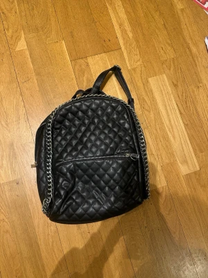 Svart ryggsäck i skinn med kedja - Snygg svart ryggsäck från Tiamo i quiltad design med kedjedetaljer och dragkedja framtill. Väskan är gjord i skinnimitation och har justerbara axelband. Perfekt för dig som vill ha en trendig och praktisk väska med plats för allt du behöver. Lite mindre i modellen. 