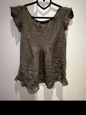 Mörkgrön / khakigrön blus med volang från MNG XS - Supersöt khakigrön / mörkgrön blus från MNG i storlek S (passar XS också). Blusen har korta volangärmar, rund halsringning och ett subtilt randigt mönster. Nedtill finns dekorativa volanger och sydda detaljer som ger en romantisk vibe. Perfekt att styla med jeans eller kjol.