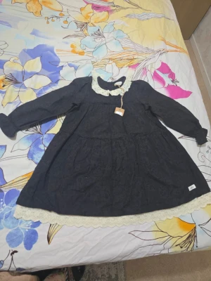 Mörkblå klänning med spets från Newbie - Supersöt mörkblå kortklänning från Newbie med långärmad design. Klänningen har en dekorativ vit spetskrage och matchande spetsdetaljer längst ner. Tyg med diskret struktur och mönster ger en vintagekänsla. Perfekt för dig som gillar romantiska detaljer.