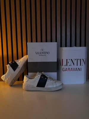 Valentino Garavani vita sneakers - Säljer ett par vita Valentino Garavani sneakers med svart band över sidan och coola nitar runt hälen. Skorna har snörning och är tillverkade i skinn. Köpta för 1500kronor OBS säljes billigt då jag inte har äktehetsbevis