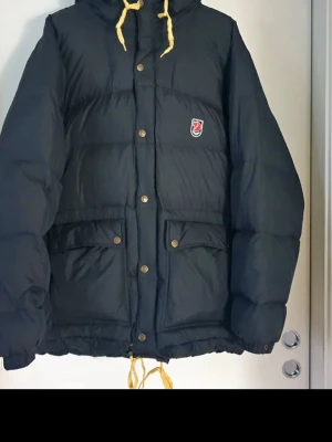 Fjällräven Expedition Lite Herr - Svart dunjacka från Fjällräven med gul snörning i huvan och flera fickor med tryckknappar. Jackan har broderad logga på bröstet och är tillverkad i polyester med dunfyllning. Perfekt för kalla dagar och har en loose passform. Dragkedjan är utbytt till silverfärgade ringar då orginal kedjan lossade. Orginalkedjan medföljer!