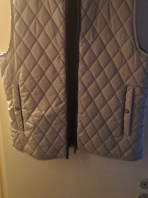 Beige quiltad dunväst med dragkedja - Quiltad beige dunväst med snyggt rutmönster och svarta detaljer vid fickorna. Västen har dragkedja framtill och två fickor med knappstängning. Insidan är svart och materialet känns mjukt och lätt. Perfekt för lager-på-lager under kyliga dagar.