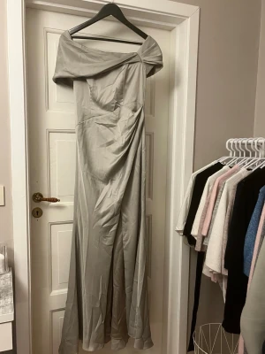 Balklänning - Elegant ljusgrå/mintgrön långklänning med offshoulder-ringning och draperad detalj framtill. Klänningen har en figurnära passform upptill och faller snyggt längs kroppen. Tillverkad i ett glansigt material som ger en lyxig känsla. Klänningen är använd fåtal gånger men fått några små trådar då det är ett känsligt material💕men absolut inget ja la märke till när jag använde den. På insidan (se bil) är de större tråd, pga nål då den va lite för öppen för mig. Passade perfekt på mig då ja är ca 1,63💕
