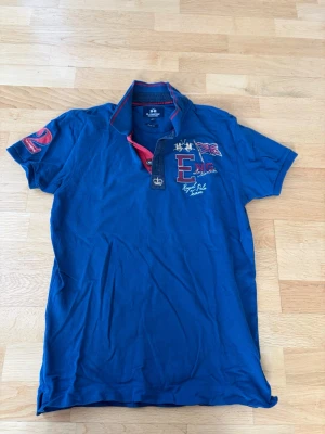 La Martina blå pikétröja, storlek M - Blå La Martina pikétröja med broderade detaljer på bröstet och ärmen. Kragen har en röd detalj.