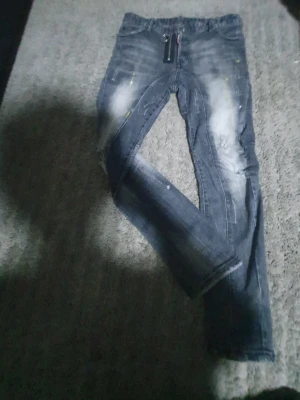 Dsquared2 blå jeans med dragkedjedetalj - Säljer ett par blå jeans från Dsquared2 med coola slitningar, ljusa tvättade partier och små gula färgstänk. Jeansen har en unik dragkedjedetalj framtill och klassiska bakfickor. Perfekt för dig som vill sticka ut lite extra.