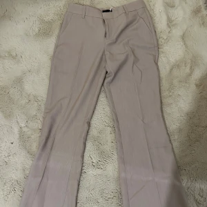 Beige kostymbyxor från Gina Tricot - Snygga beige kostymbyxor från Gina Tricot i rak modell. Byxorna har hällor för bälte, diskreta sidofickor och stängs med dragkedja och knapp. Perfekta för en clean och stilren look. Materialet är mjukt och har en lätt struktur.