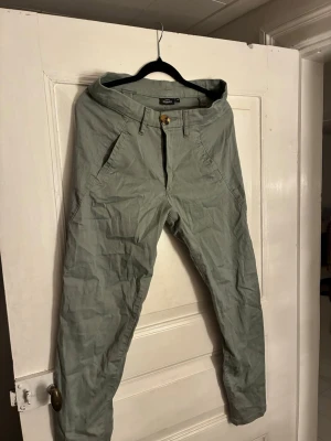 Gröna chinos från KappAhl, storlek 38 - Snygga ljusgröna chinos från KappAhl i storlek 38. Byxorna har slim passform, raka ben och klassiska fickor fram och bak. Tillverkade i bomull med bälteshällor och stängs med knapp och dragkedja. Perfekta till en avslappnad stil.