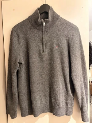 Grå stickad half zip tröja GANT - Mysig grå stickad tröja från GANT med half zip och hög krage. Klassisk broderad logga på bröstet. Perfekt till kyliga dagar och enkel att matcha med jeans eller chinos.