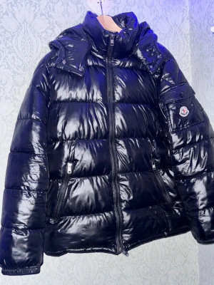 Svart Moncler pufferjacka - Säljer en svart, glansig pufferjacka från Moncler med hög krage och avtagbar huva. Jackan har dragkedja framtill, två sidofickor med dragkedja och en ficka på ärmen med Moncler-logga. Insidan har ett unikt serietidningsmönster. Perfekt för kalla dagar.