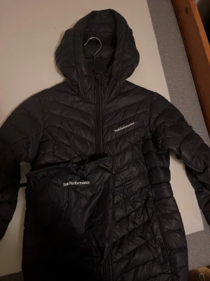 Svart dunjacka Peak Performance - Säljer en svart dunjacka från Peak Performance med huva och dragkedja framtill. Jackan har ett stilrent, quiltat mönster och logga på bröstet. Perfekt för kalla dagar och enkel att packa ner i medföljande påse. Långärmad och lättviktig design.