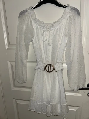 Vit kortklänning med bälte, perfekt för student!  - Säljer en vit kortklänning med långärmade puffärmar och ett snyggt guldigt bälte i midjan. Klänningen har ett diskret prickigt mönster, volang nertill och knytning vid bröstet. Materialet känns lätt och luftigt, perfekt för varmare dagar. Lappen är borta och har använt 1 gång