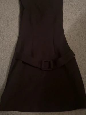 Brun kortklänning med bälte - Säljer en brun kortklänning med markerad midja och matchande bälte. Klänningen har en enkel, stilren design utan mönster och är lätt utsvängd nedtill. Perfekt för dig som gillar minimalistisk stil och vill ha något klassiskt i garderoben.