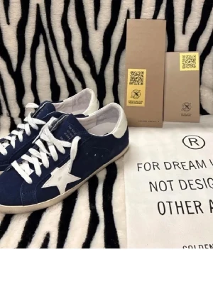 Golden Goose blå sneakers med vit stjärna - Säljer ett par snygga Golden Goose sneakers i marinblå mocka med vit stjärndetalj på sidan och vit häl. Skorna har rund tå, platt sula och vita snören. Märket syns tydligt på hälen och tungan. Perfekta för dig som gillar coola detaljer och streetstyle. Hör av er vid frågor✅