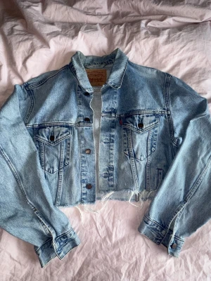 Croppad jeansjacka från Levi's - Säljer en ljusblå croppad jeansjacka från Levi's med rå kant nertill. Klassisk modell med två bröstfickor med knappar och långa ärmar. Perfekt till lager på lager och ger en chill vibe till din outfit. Liten fläck på ena armen.