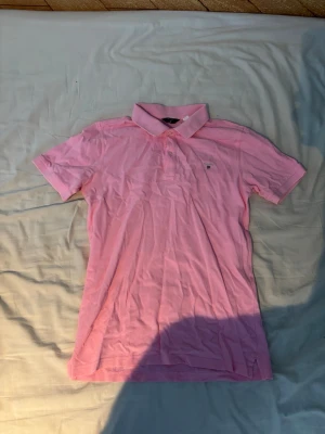 Rosa pikétröja från Gant - Säljer en klassisk rosa pikétröja från Gant med korta ärmar och krage. Tröjan har knappar framtill och Gant-logga broderad på bröstet. Tillverkad i mjuk bomull som känns skön mot huden. Perfekt för en avslappnad och stilren look.