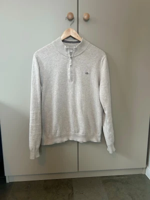 Grå half zip tröja från GANT - Snygg beige tröja från GANT med half zip och ribbade muddar. Tröjan har en hög krage med marinblå ränder och diskret GANT-logo broderad på bröstet. Perfekt till chill vardagsstil och funkar året runt. Är i mycket fint skick och är inte använd allt för mycket. Köpt för 1000