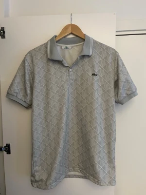 Grå piké från Lacoste - Säljer en grå pikétröja från Lacoste med diskret geometriskt mönster i vitt. Storleken är L men passar mera som M. Pris kan diskuteras vid snabb affär